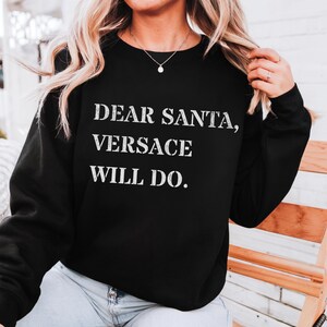 Puede incluir: Sudadera negra con el texto blanco "DEAR SANTA, VERSACE WILL DO.". La sudadera es de cuello redondo y la lleva una persona con cabello rubio.