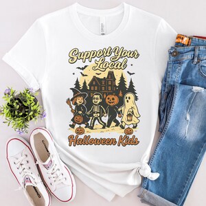 May include: White t-shirt with a Halloween-themed graphic. The design features a haunted house, bats, and children in costumes with the text "Support Your Local Halloween Kids". The shirt is paired with blue jeans and white sneakers.