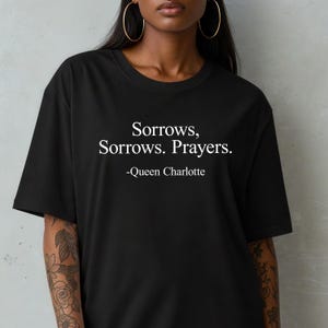 May include: Black t-shirt with the quote "Sorrows, Sorrows. Prayers." attributed to Queen Charlotte in white text. The shirt has a crew neck and short sleeves.