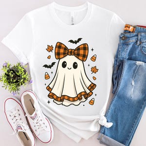 Może przedstawiać: Biała koszulka z kreskówkowym duchem z kratkowaną kokardą, otoczonym nietoperzami, jesiennymi liśćmi i cukierkami kukurydzianymi. Koszulka jest połączona z niebieskimi dżinsami i białymi trampkami, tworząc swobodny, świąteczny wygląd na Halloween.