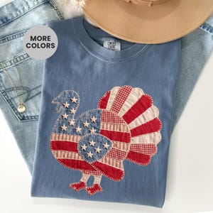 Puede incluir: Una camiseta azul de cuello redondo con una gran aplicación de pavo en la parte delantera. El pavo está diseñado con un patrón patriótico rojo, blanco y azul, que incluye estrellas y rayas de tela. La camiseta tiene una etiqueta Comfort Colors.