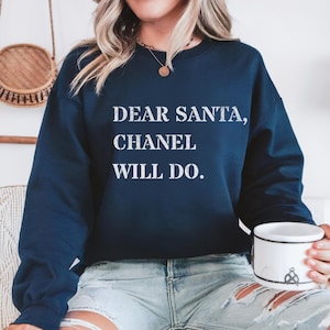 May include: A navy blue crew neck sweatshirt featuring the white text "DEAR SANTA, CHANEL WILL DO." The sweatshirt is being worn by a person. The person is holding a white mug with a design on it.