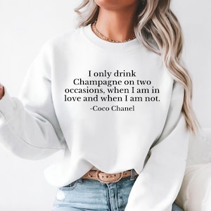 May include: White sweatshirt featuring the quote "I only drink Champagne on two occasions, when I am in love and when I am not. -Coco Chanel" in black text. The model is wearing blue jeans and a gold necklace.