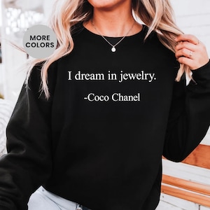 May include: Black sweatshirt featuring the quote "I dream in jewelry." attributed to Coco Chanel in white text. The text is centered on the front. A circular button in the upper left corner reads "MORE COLORS".