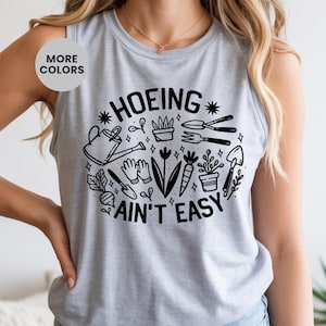 Puede incluir: Una camiseta sin mangas gris claro con un gráfico negro que dice "HOEING AIN'T EASY". El gráfico presenta herramientas de jardinería, plantas y una regadera dispuestas en un diseño circular. La camiseta sin mangas es sin mangas.