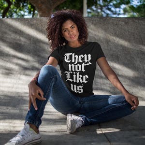 May include: A person wearing a black t-shirt with the text "They not like us" in white gothic lettering. The person is sitting on a concrete surface and wearing blue jeans and white sneakers.