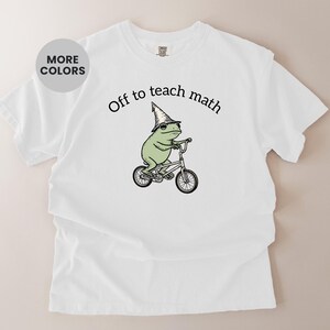 May include: White t-shirt featuring a cartoon frog in a wizard hat riding a bicycle, with the words "Off to teach math" above. The shirt has a crew neck and short sleeves. The frog is green and the text is black.