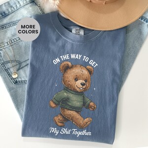Puede incluir: Camiseta azul con un oso de peluche marrón con un suéter verde. El texto "ON THE WAY TO GET" está encima del oso, y "My Shit Together" debajo. La camiseta está sobre un fondo de mezclilla.