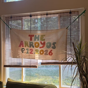 Könnte beinhalten: Ein rechteckiges Banner aus cremefarbenem Stoff mit bunten, gemusterten Stoffbuchstaben, die den Schriftzug THE ARROYOS und das Datum 9.12.2026 bilden. Das Banner ist an einer grünen Schnur vor einem Fenster befestigt und zeigt diverse Texturen.
