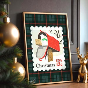May include: Framed Christmas-themed artwork featuring a vintage postage stamp design. The stamp depicts a red mailbox with a snow-covered roof, a package, and the text "Christmas 13c USA." The frame has a green and red plaid pattern.