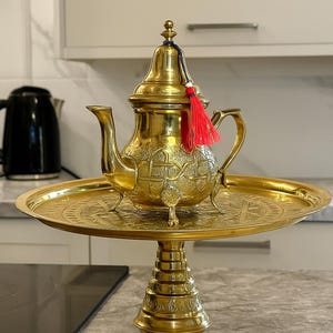 Könnte beinhalten: Eine goldfarbene Teekanne mit aufwendigen Mustern und einer roten Quaste, platziert auf einem passenden dekorativen Tablett und Ständer. Im Hintergrund ist ein schwarzer Wasserkocher zu sehen. Die Teekanne und das Tablett sind aus Metall.