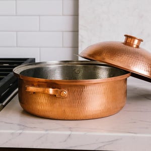 May include: A copper cooking pot with a matching lid, set on a marble countertop next to a gas hob. The pot has a hammered texture and a shiny, metallic finish. The lid is slightly ajar, revealing the pot's interior.
