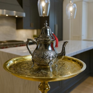 Könnte beinhalten: Eine silberne Teekanne mit aufwendigen Designs und einer roten Quaste steht auf einem goldfarbenen Tablett. Das Tablett wird von einem goldenen Ständer getragen. Die Küche im Hintergrund hat dunkle Schränke und eine Marmorarbeitsplatte.