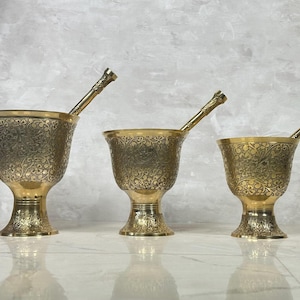 Könnte beinhalten: Drei verzierte, goldfarbene Mörser und Stößel-Sets. Jeder Mörser hat einen ausgestellten Rand und einen Sockel, mit aufwendigen floralen Mustern, die über die Oberfläche geätzt sind. Die Stößel sind goldfarben und haben strukturierte Griffe.