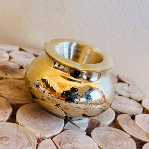May include: A gold hammered metal ashtray with a round body and a wide, shallow opening. The ashtray is sitting on a surface of stacked wooden rounds.