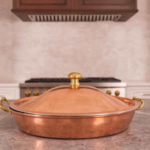 以下が含まれることがあります： 蓋と金色の取っ手とつまみが付いた銅製の調理鍋。鍋はキッチンの灰色のカウンタートップに置かれています。鍋は丸く、光沢のある仕上がりです。