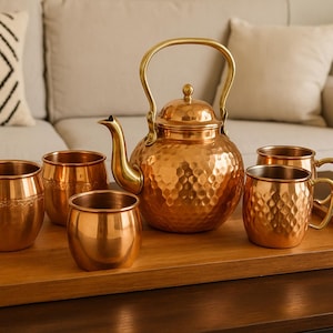 May include: A copper tea set, including a hammered kettle with a curved spout and handle, and six matching mugs and cups, arranged on a wooden tray. The set is displayed on a dark brown table in front of a beige sofa.