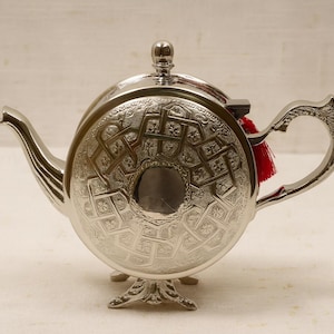 Puede incluir: Una tetera plateada con intrincados patrones geométricos. La tetera tiene un cuerpo redondo, un pico curvo y un asa decorativa. Una pequeña borla roja cuelga de la tapa. La tetera se apoya sobre una base decorativa.