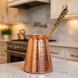 Op de afbeelding: Een koperkleurige Turkse koffiepot met een gehamerde textuur en een goudkleurige handgreep. De pot heeft een brede basis die naar boven taps toeloopt. De handgreep heeft een geometrisch ontwerp. De pot staat op een aanrecht in een keuken.