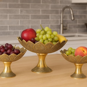 May include: Three decorative brass fruit bowls with pedestal bases. The bowls are filled with fresh fruit, including grapes, apples, and peaches. The bowls have a detailed floral design and are arranged on a wooden surface.