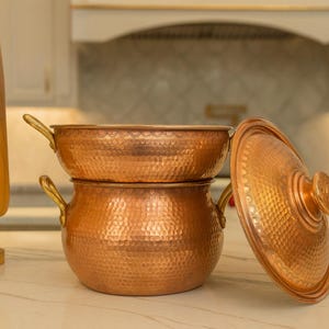 Pode incluir: Duas panelas de cozinha de cobre com textura martelada. Uma panela é empilhada em cima da outra. A panela superior é uma frigideira rasa, e a panela inferior é uma panela redonda com duas alças. Uma tampa de cobre está ao lado.