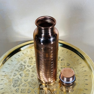 May include: A copper water bottle with a screw-on lid sits on a gold-colored tray. The bottle has a cylindrical shape with horizontal ridges. The lid is next to the bottle. The tray has an intricate pattern.