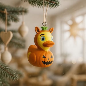 May include: A Halloween-themed Christmas tree ornament. The ornament is a duck wearing a pumpkin costume. The duck has a yellow face and orange beak. The pumpkin costume is orange with a carved jack-o'-lantern face. The ornament hangs from a beige string.