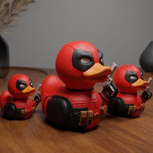 Può includere: Tre paperelle di gomma rosse e nere a forma di Deadpool, un supereroe immaginario. Le paperelle tengono pistole giocattolo e hanno una maschera nera per gli occhi.
