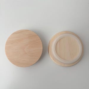 Op de afbeelding: Twee houten potdeksels. Eén is een massief, rond stuk lichtgekleurd hout. De andere heeft een heldere, cirkelvormige siliconen ring rond de rand. De deksels zijn ontworpen om een ​​container af te sluiten.