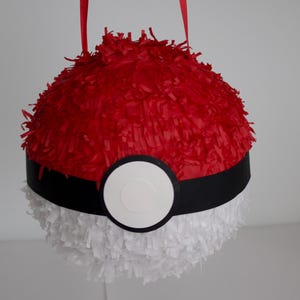 May include: A spherical piñata designed to resemble a Poké Ball, featuring a top half covered in red fringe, a bottom half covered in white fringe, and a central black horizontal band with a circular white button in the centre and red straps.