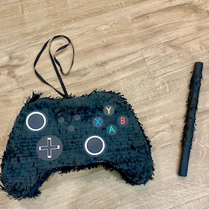 May include: A black video game controller shaped piñata covered in fringe paper with a black hanging ribbon. The controller features a white directional pad, two white circular joysticks, and four coloured buttons labelled Y, B, A, and X. A matching black stick is included.