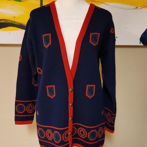 May include: Navy blue cardigan with red trim and decorative geometric and shield-like patterns. The long-sleeved sweater has a V-neck, gold buttons, and is displayed on a mannequin.
