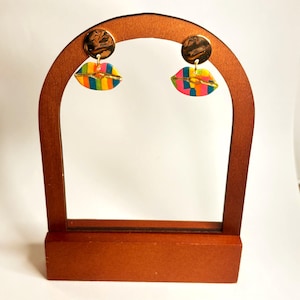 May include: A pair of colorful lip-shaped earrings with a gold-colored accent. The earrings are displayed on a wooden stand. The lips are painted with rainbow stripes and gold accents, creating a vibrant and eye-catching design.