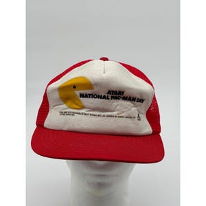 May include: Red and white Atari Pac-Man Day trucker hat. The hat features a red brim and mesh back, with a white front panel displaying the Pac-Man character and the text "ATARI NATIONAL PAC-MAN DAY".