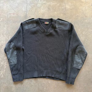 May include: A black V-neck sweater featuring a ribbed knit texture, long sleeves, and solid black fabric patches on the shoulders and elbows. The interior collar tag displays the text KM in red and yellow lettering against a black background.