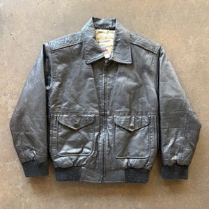 May include: A black leather jacket with a zip-up front, two front pockets, and a collared neckline. The jacket has a ribbed knit hem and cuffs. The interior lining is visible at the collar. The jacket is a vintage style.
