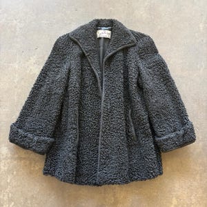 Puede incluir: Un abrigo gris oscuro texturizado con cuello con solapas y mangas con puños. El abrigo tiene una forma ligeramente acampanada y un diseño de un solo pecho. La tela parece ser un material denso, posiblemente lana o piel sintética.