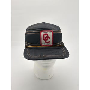 May include: Black cap with gold stitching and a rectangular patch. The patch reads "STOP AT" above a red and white logo with the letters "GC" and "TARRAS, PA" below.