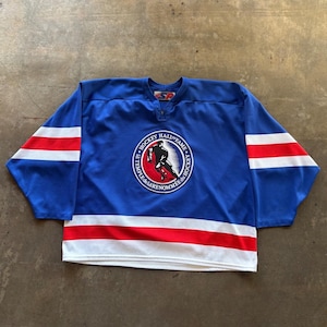 May include: A blue hockey jersey featuring a circular Hockey Hall of Fame logo in the center with a red background and a black silhouette of a hockey player. The sleeves and bottom hem have horizontal white and red stripes. A small tag is at the neck.