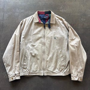 May include: Tan Nautica jacket with a full zipper, two front pockets, and buttoned cuffs. The collar lining features red, green, and blue stripes. A small Nautica logo is embroidered on the chest.