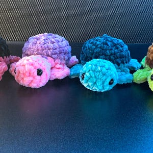 May include: Six crocheted turtle toys in a row, each with a soft, textured appearance. The turtles are in shades of pink, purple, teal, brown, green, and black. Each turtle has black safety eyes and flippers that match the body colour.