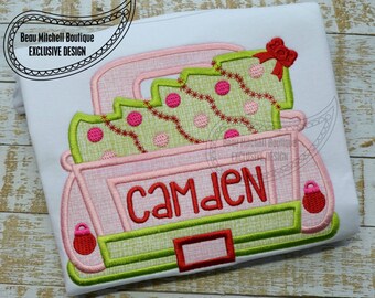 May include: Pink and green embroidered design of a pickup truck carrying a Christmas tree with the name Camden on the back. The truck has a red bow on top of the tree and red ornaments on the back.