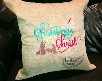 May include: A square, beige pillow with the embroidered text "Christmas begins with Christ" in teal and pink. A silhouette of the nativity scene is below the text. The pillow has a textured appearance and is likely made of linen or a similar fabric.
