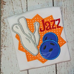 May include: A white t-shirt with an orange and white starburst design. The design features a bowling pin, two bowling balls, and the word "Jazz".