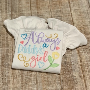 Puede incluir: Camiseta blanca con mangas abullonadas con el texto bordado "Always a Daddy's girl" en escritura colorida con corazones y una flor.
