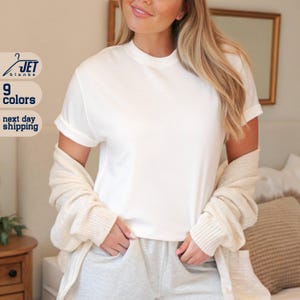May include: A white short-sleeved t-shirt with a rolled-up sleeve. The shirt is paired with gray sweatpants and a cream-colored cardigan. The text "JET blanks" and "9 colors" are visible in the image.
