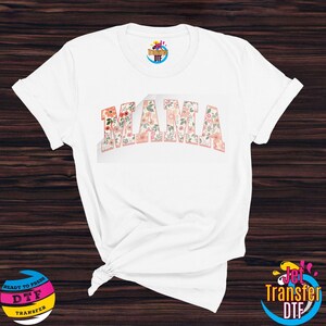 May include: White t-shirt with the word "MAMA" in pink letters with a floral pattern. The floral pattern is a mix of pink, orange, and yellow flowers.