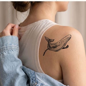May include: A black and white whale temporary tattoo design. The whale is depicted in a detailed, artistic style, with visible lines and shading. The tattoo is placed on a person's shoulder.