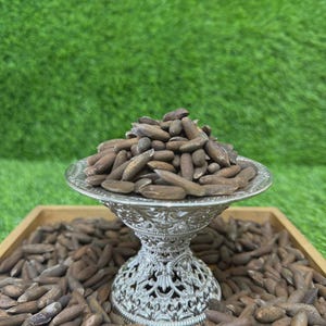 May include: A silver decorative bowl filled with brown pine nuts, set on a wooden tray. The bowl has an ornate, detailed design. The background is a green, grassy surface.