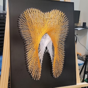 Könnte beinhalten: Ein String-Art-Stück, das einen Zahn darstellt. Das Kunstwerk zeigt orangefarbenen und weißen Faden, der sorgfältig um Nägel auf einem schwarzen Hintergrund gewebt ist. Das Zahndesign wird auf einer Holzstaffelei angezeigt.